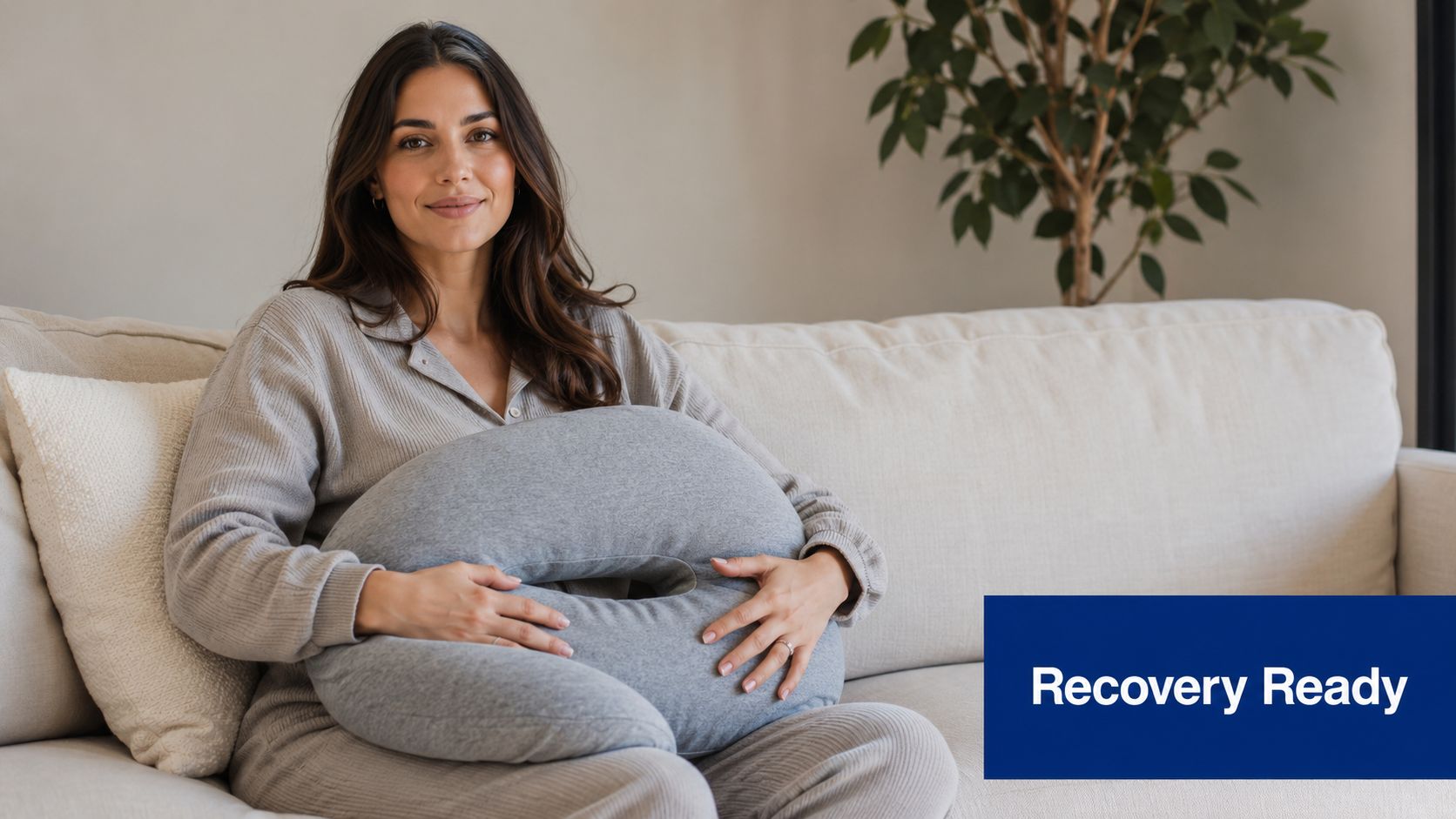 A woman smiling while holding a grey, crescent-shaped recovery pillow designed for post-surgery comfort on a sofa.